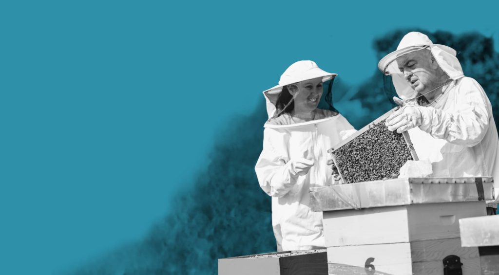 two people in bee suits standing next to a beehive.
